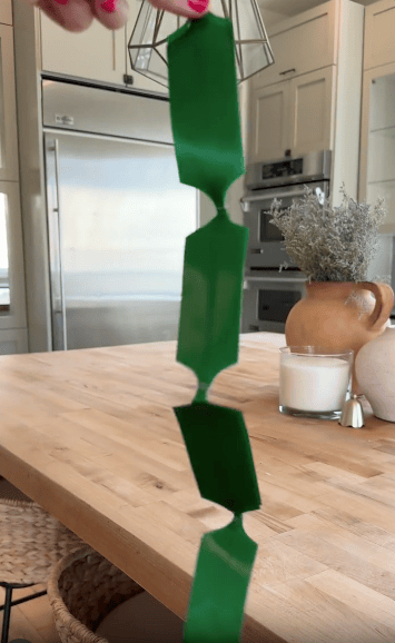 A hand holds a green paper strip with cut-out sections, forming a folding chain shape, over a wooden kitchen table with decor and a stainless-steel fridge in the background.