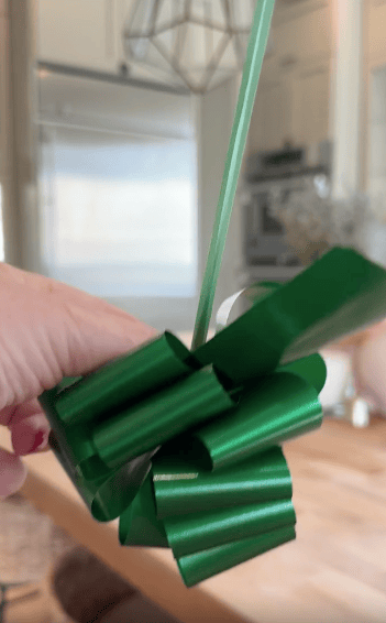 A hand with red nail polish holds a shiny green gift bow with attached curling ribbon, in a bright kitchen setting.