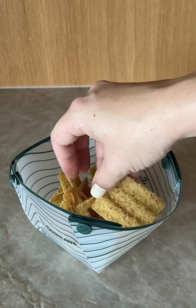 A hand reaches into a small, triangular, collapsible silicone bowl filled with ridged potato chips, placed on a gray countertop with a wooden background.