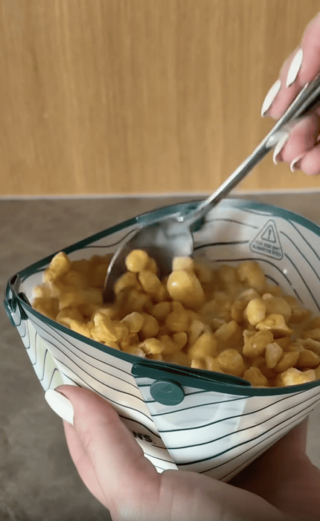 A hand holds a measuring cup filled with corn kernels while another hand stirs them with a spoon. The measuring cup is transparent with measurement markings and has a warning symbol visible.