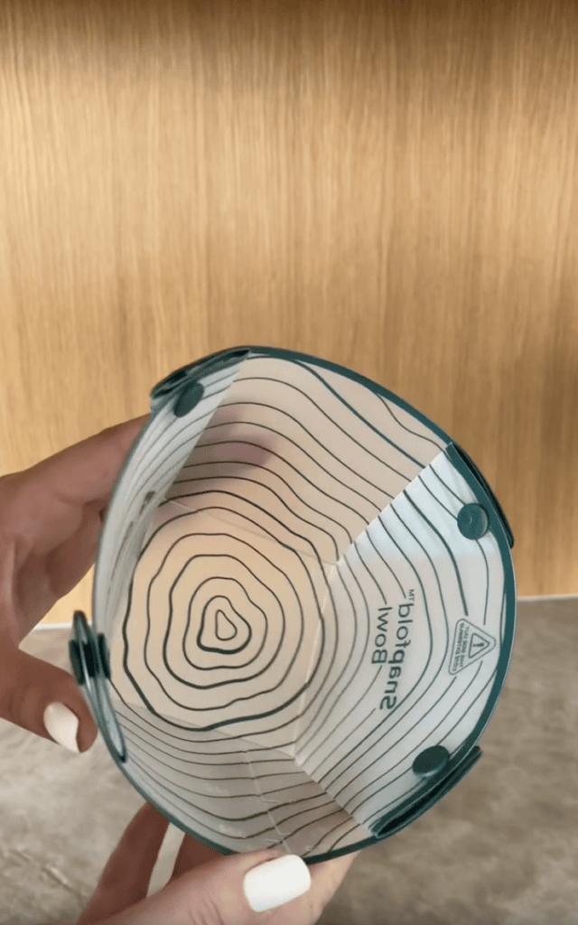 A person holds a clear, collapsible bowl with concentric circle designs, teal edges, and a wood background. The bowl is labeled “smatold bowl” and has symbols printed inside.