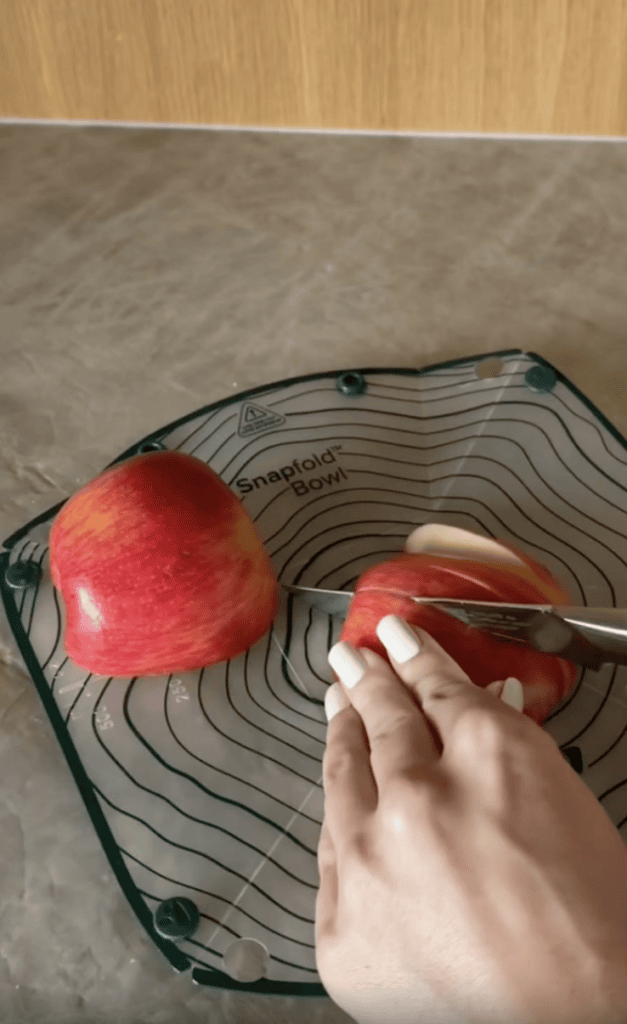A hand with white nail polish cuts a red apple on a green Snapfold Bowl mat placed on a gray countertop.