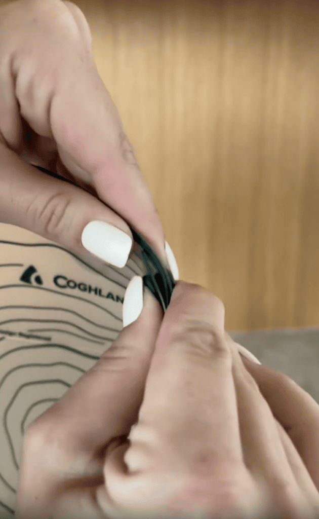 Close-up of hands with white nail polish sealing a dark green bag. The background shows part of a patterned surface with the Coghlan’s logo and a wooden texture.