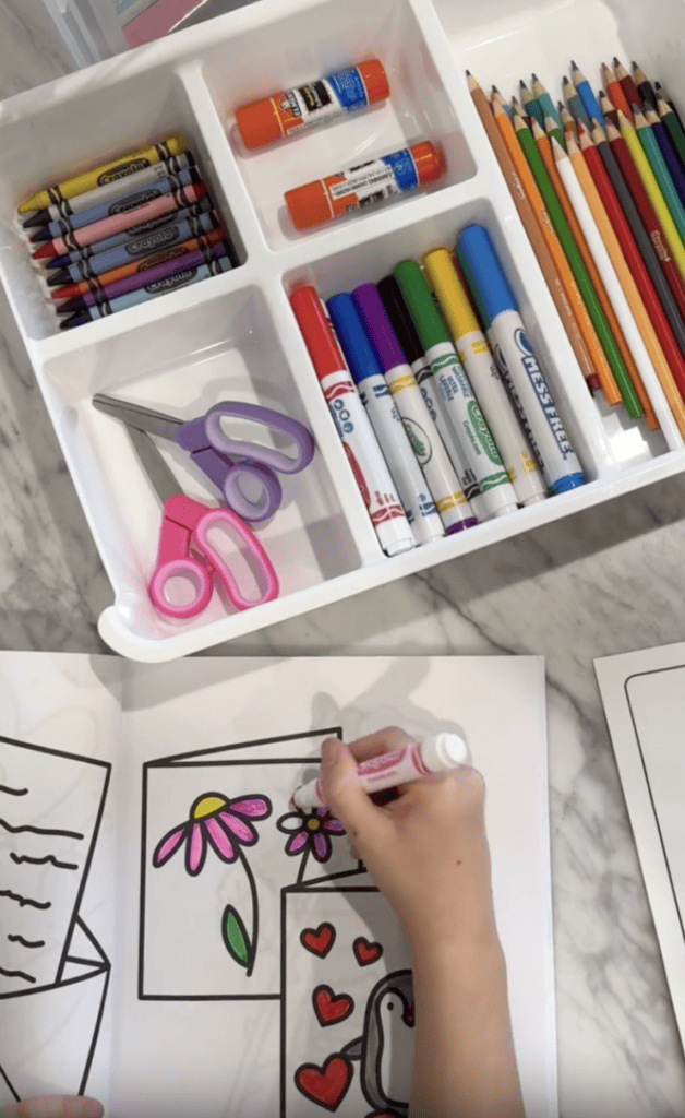 A tray holds crayons, glue sticks, colored pencils, scissors, and markers. Below, a child colors a drawing of flowers, hearts, and a penguin with a pink marker on white paper.