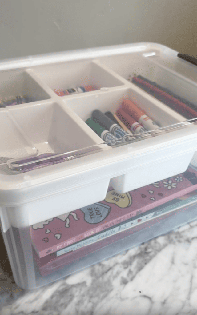 A clear plastic organizer holds markers, pencils, glue sticks, and a pink activity book labeled “My First Book of Valentines” on a marble surface.