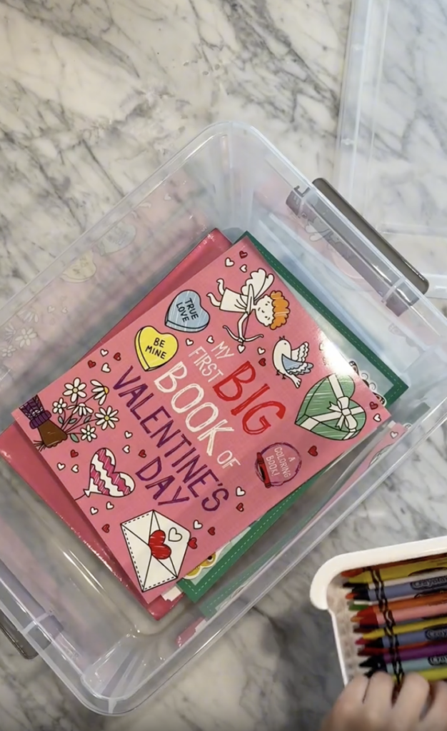 A plastic bin holds a pink Valentine’s Day book with cartoons, hearts, and flowers on the cover. Next to it is a small container filled with colorful crayons. The bin sits on a marble surface.