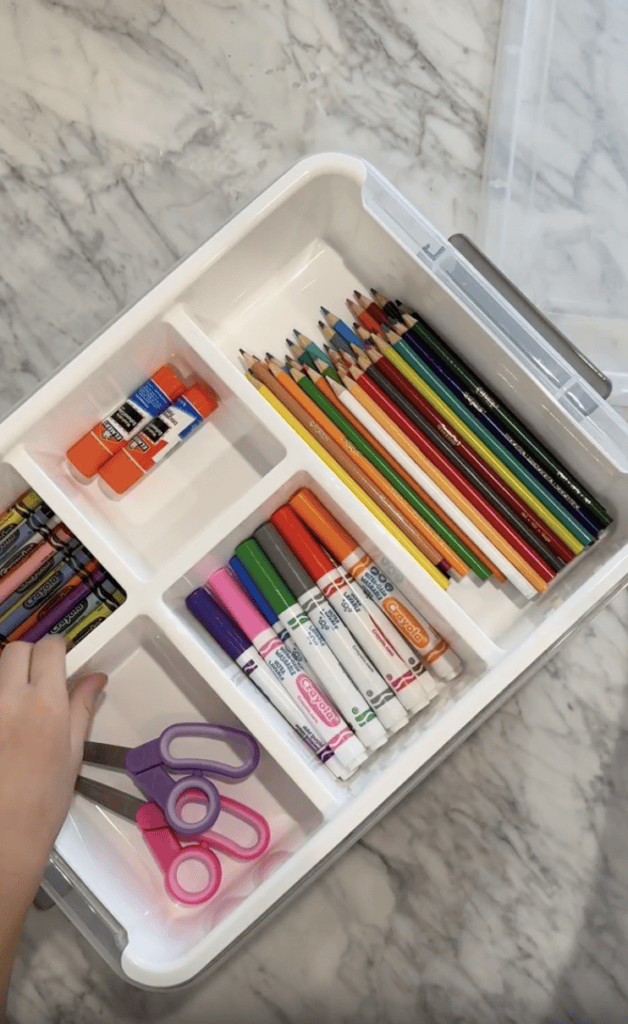 A hand reaches for purple scissors in a white organizer tray holding colored pencils, crayons, markers, glue sticks, and scissors on a marble surface.