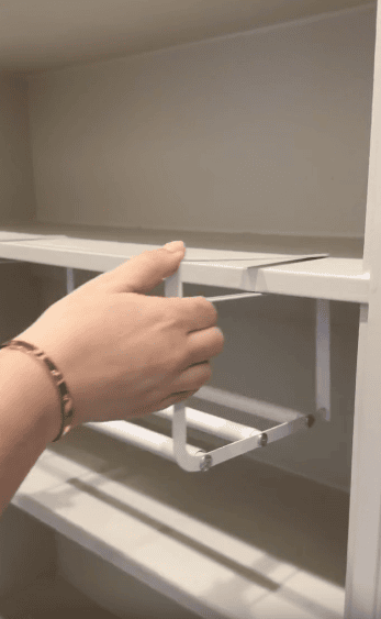 A person attaches a white metal hanging rack under a shelf in a cabinet, wearing a gold bracelet. The rack is designed for organizing items and saving space within the shelving unit.