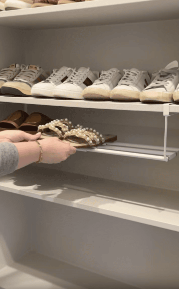 A person places a pair of pearl-studded sandals on a shoe rack in a closet, with rows of sneakers and other footwear visible on the shelves above.