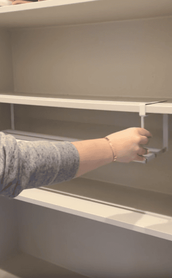 A person in a gray sweater installs a white shelf divider onto a white shelf in a closet or storage area.