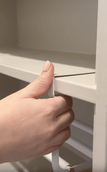 A hand pulls a white plastic tab along the edge of a beige shelf, appearing to remove adhesive tape or a protective strip from the shelf’s surface.