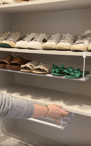 A person adjusts a white shelf in a closet. Above, neatly arranged shoes include sneakers, brown sandals, pearl-decorated sandals, and green bow sandals on a higher shelf.