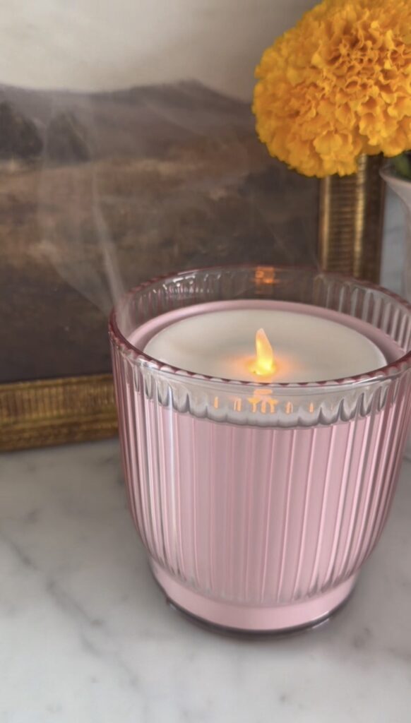 A pink, ribbed glass candle sits on a marble surface, with a soft flame. In the background, there’s a yellow flower and a brown picture frame. Wisps of smoke are visible above the candle.