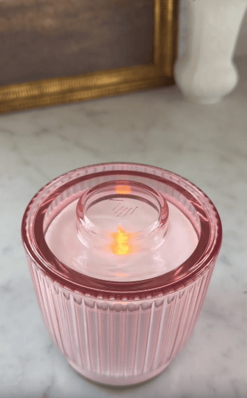 A pink ribbed glass candle sits on a marble surface, glowing with a lit flame inside. A gold-framed picture and a white vase are blurred in the background.