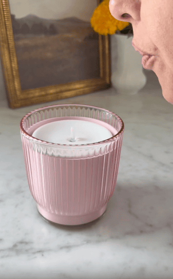 A person blowing to extinguish a lit candle in a pink ribbed glass holder on a marble surface; framed art and flowers in the background.
