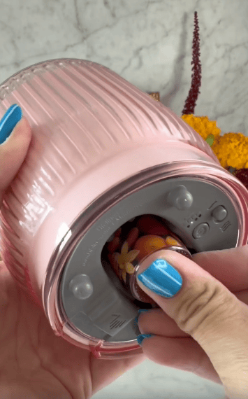 A person with blue nail polish inserts a floral-scented cartridge into the bottom of a pink, ribbed air freshener device, with yellow flowers blurred in the background.
