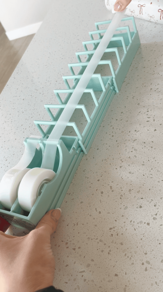 A hand holds a teal dispenser with two rolls of tape and several compartments, placed on a speckled countertop. Another hand pulls a strip of tape from the dispenser.