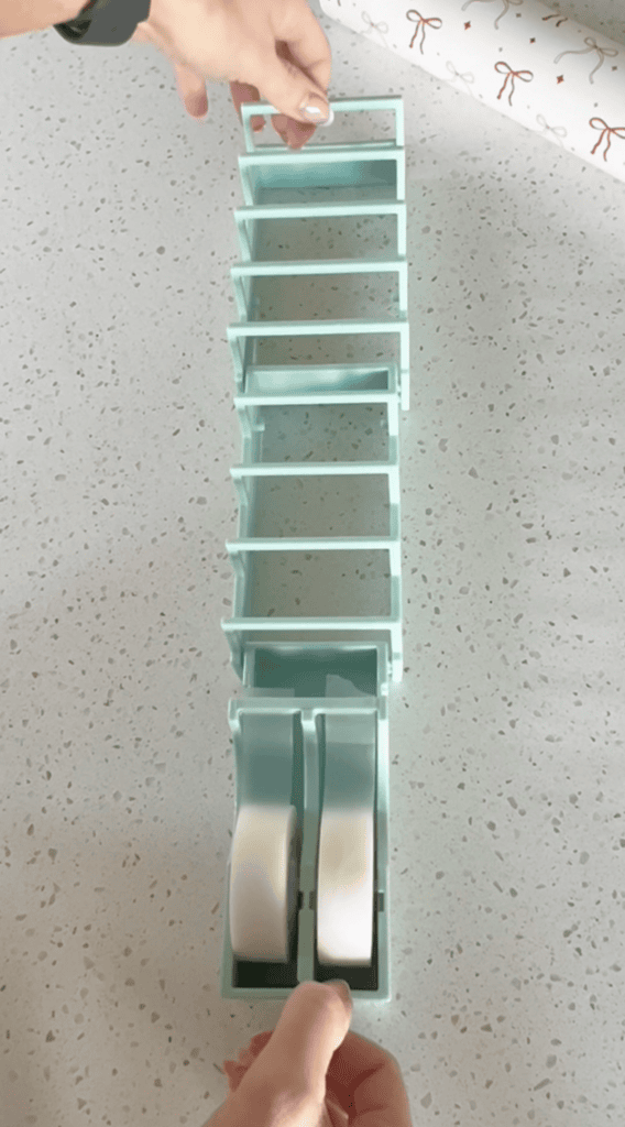 A hand places a small object into a mint green organizer holding two tape rolls at the bottom, on a speckled countertop. Wrapping paper is partially visible at the top right corner.