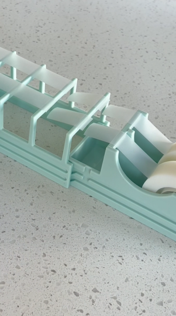 A mint green plastic organizer holds two rolls of white tape on a speckled countertop. The organizer has several empty compartments and a slot for dispensing tape.
