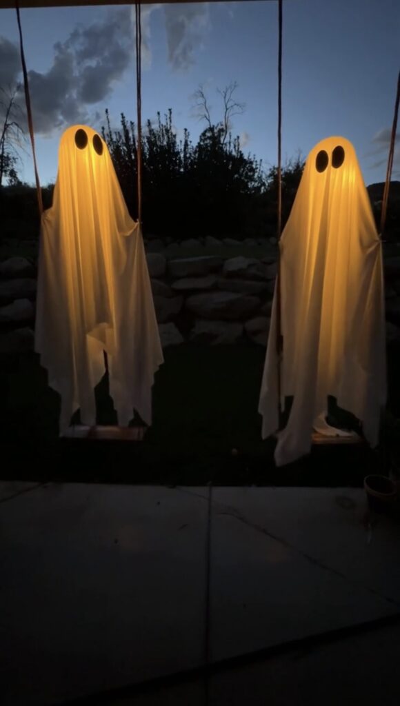 Two ghost decorations made from white sheets with glowing lights and black eyes hang on swings outdoors at dusk.