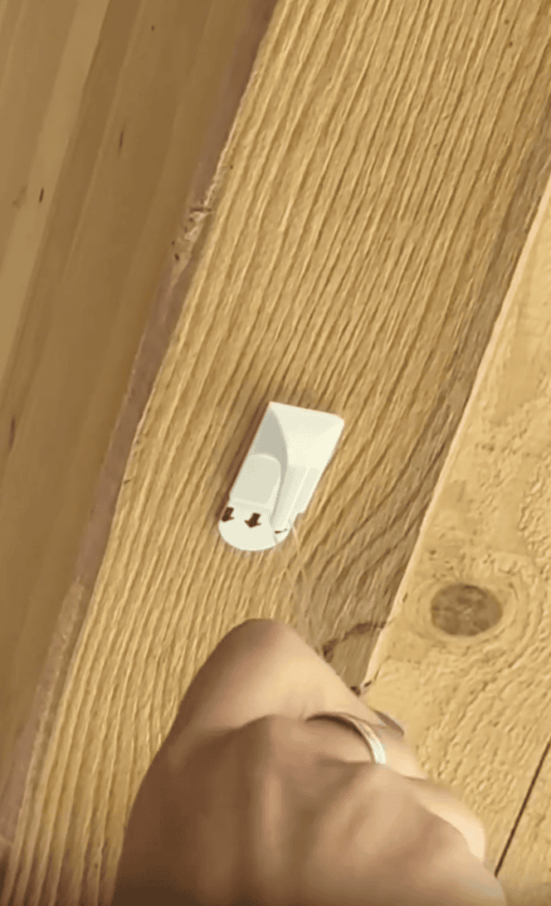 A hand holds a tool near a white cable clip attached to a wooden surface. The clip has two arrows pointing down, indicating where to press or insert something.