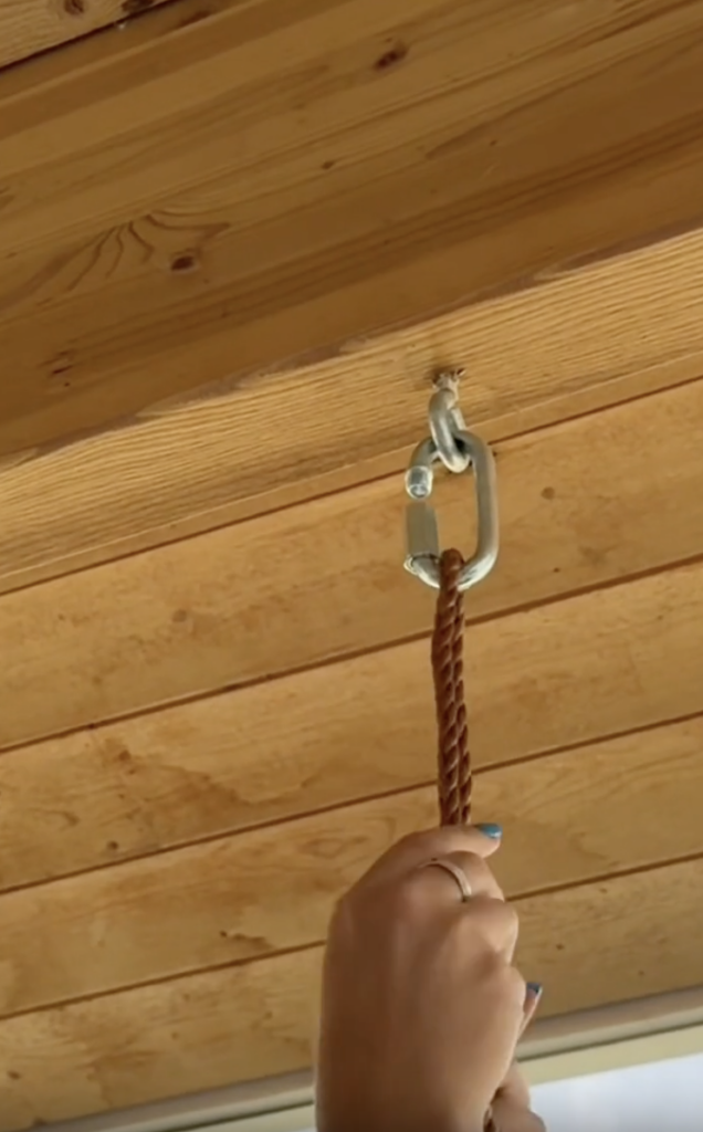 A hand holds a brown rope that is attached by a metal carabiner to a hook on a wooden ceiling.