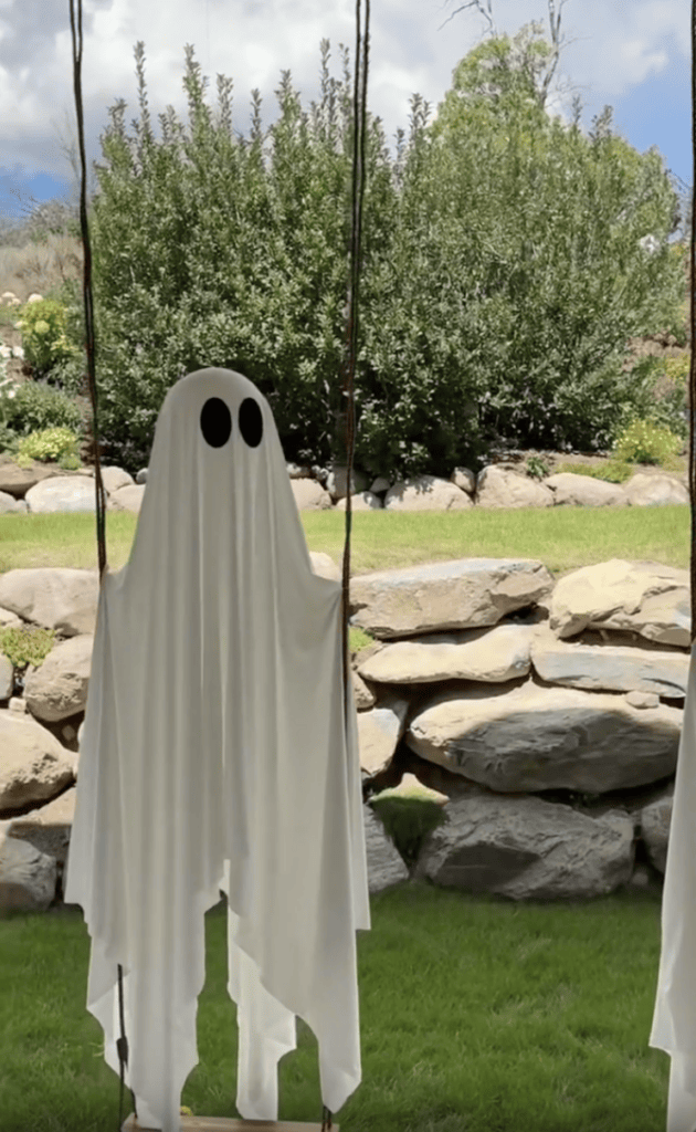 A Halloween ghost decoration made from a white sheet with black eyes hangs outdoors in a green yard with stone walls and bushes in the background.