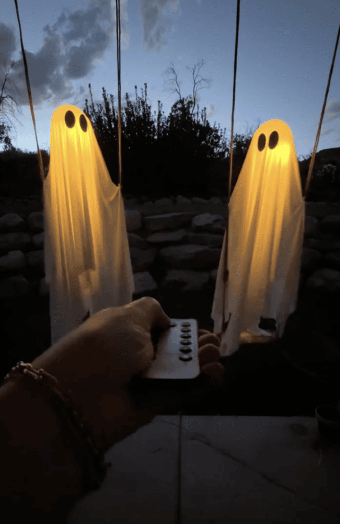 A hand holds a remote toward two glowing ghost decorations hanging outdoors at dusk, with rocks and trees in the background.