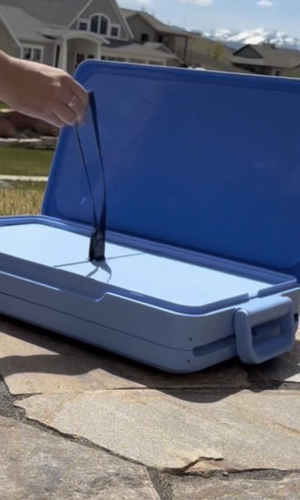 A person opens a large blue plastic case outdoors, holding a dark strap attached inside the case. Houses and mountains are visible in the background on a sunny day.