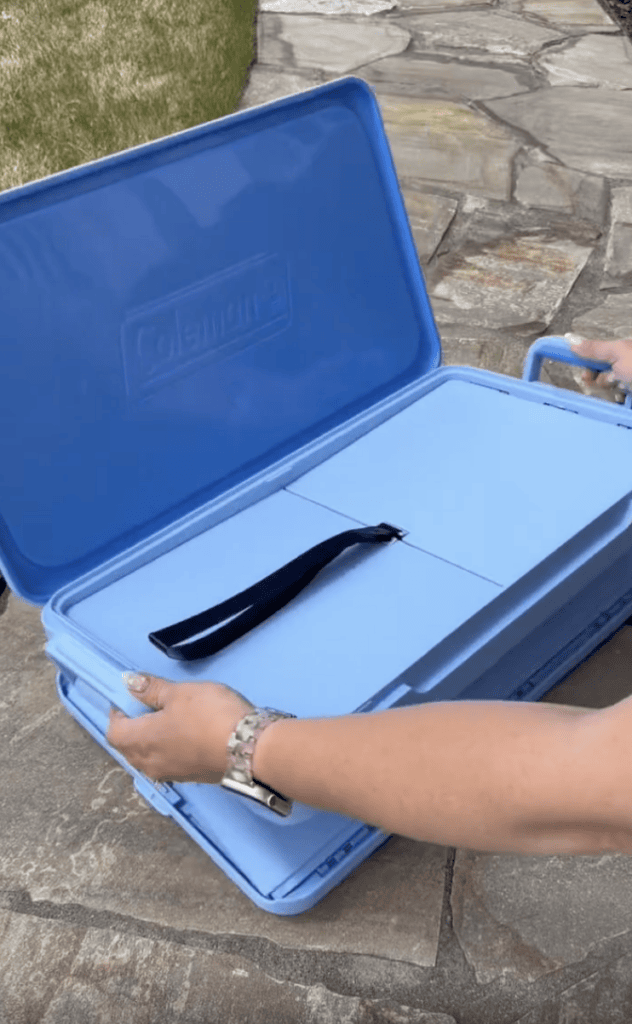 A person opens a blue Coleman carrying case on a stone path, revealing a folded portable table with a black handle inside.