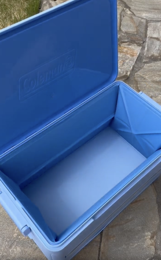 An open, empty blue Coleman cooler sits on a stone patio with grass visible in the upper left corner.