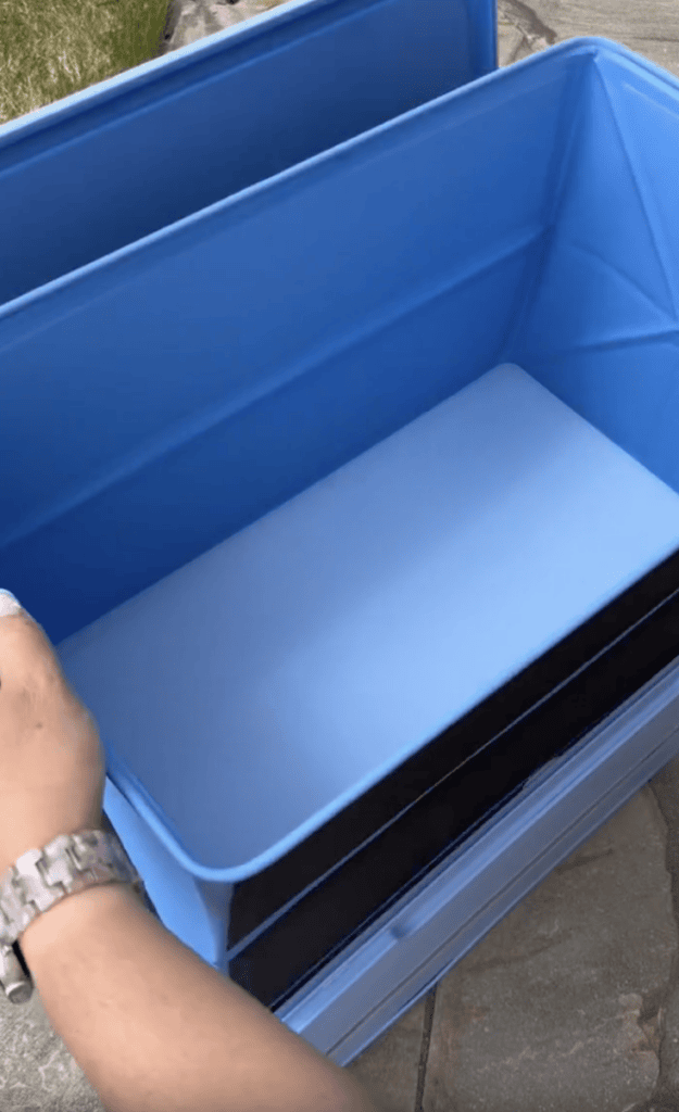 A person’s hand holding open a blue plastic storage bin with a white rectangular object inside, placed on a stone pavement outdoors.