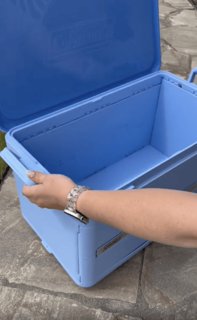 A person opens a large blue Coleman cooler outdoors on a stone surface. The cooler is empty and the lid is lifted.