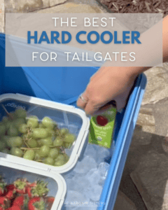 A hand places a juice box in a blue hard cooler filled with ice, grapes, and strawberries. Text reads: “The Best Hard Cooler for Tailgates.”.