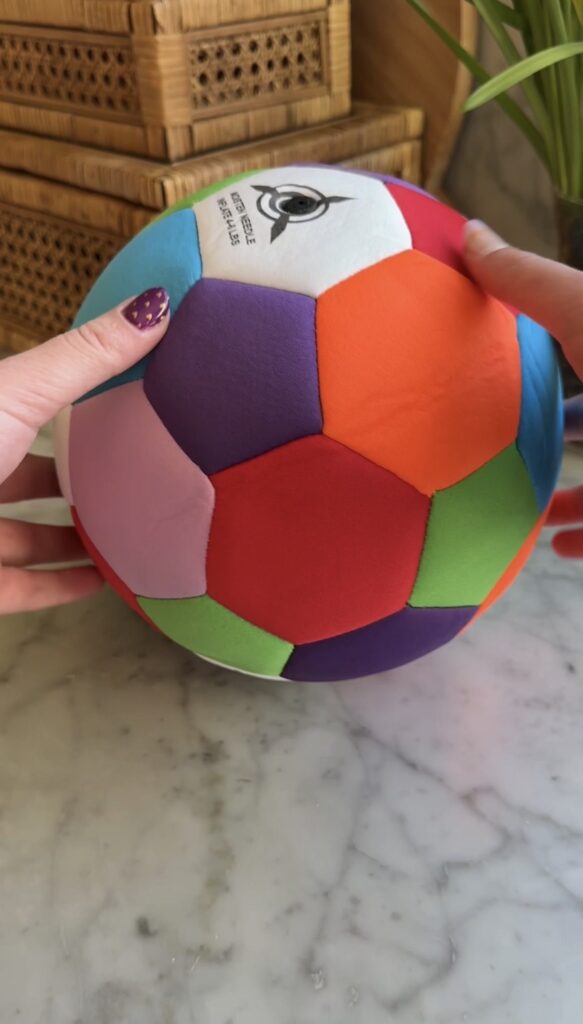 Two hands hold a colorful soccer ball made of fabric patches in red, orange, green, purple, pink, white, and blue on a marble surface. One hand has purple nail polish.