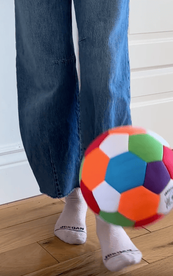 A person wearing blue jeans and white socks kicks a colorful soccer ball indoors on a wooden floor.