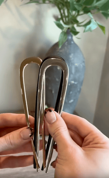 Two hands hold three metallic U-shaped hairpins—two gold, one silver—against a blurred background with a dark vase and green leafy plant.