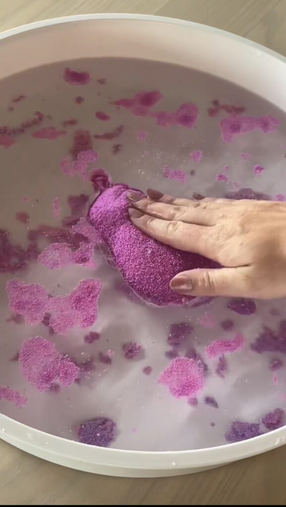 A hand presses a purple, sand-like object in a white bowl filled with water, causing the object to dissolve and break into smaller pieces.