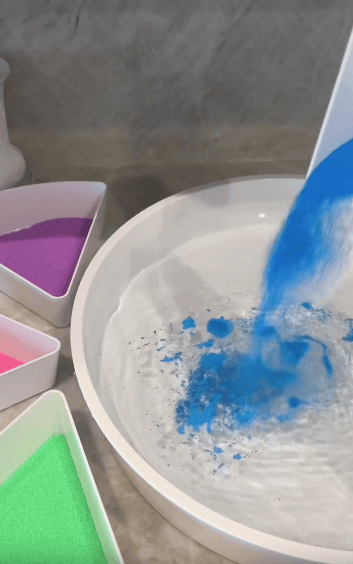 Blue powder being poured into a bowl of water, with containers of pink, purple, and green powder nearby on a countertop.