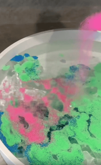 A white bowl of water with vibrant green, blue, and pink powder sprinkled on the surface, creating colorful patterns as the powder spreads.