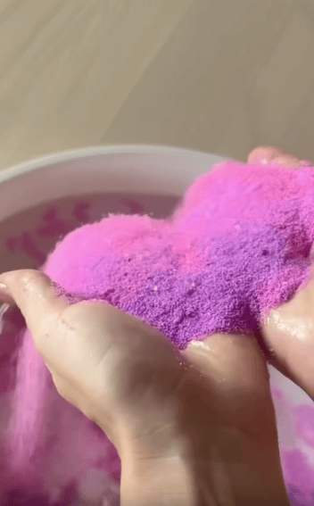 Two hands hold wet, clumpy sand with a gradient of pink and purple colors over a white bowl.