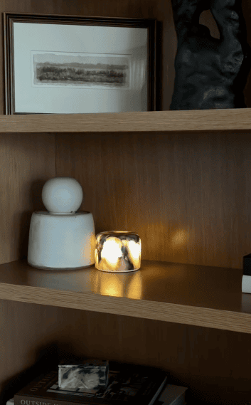 A small marble-patterned candle holder glows on a wooden shelf beside a white abstract sculpture and stacked books.