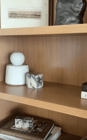 Wooden shelves display a white orb atop a vase, a marbled candle, a black rectangular object, and stacked books, including one with the visible title “OUTSIDE IN.”.