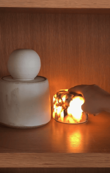 A hand touches a glowing, marbled lamp on a wooden shelf next to a white, round-lidded ceramic jar.