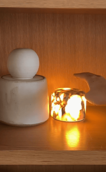 A white ceramic container and a lit marble-patterned candle sit on a wooden shelf. A hand reaches toward the glowing candle, casting a warm light against the wood background.