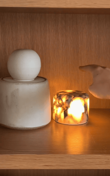 A hand reaches toward a small, glowing marble-patterned candle next to a round white ceramic container on a wooden shelf.