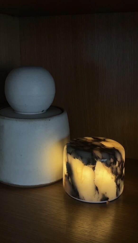 A marble-patterned candle glows beside a white, round-lidded ceramic container on a wooden surface, creating a warm and cozy ambiance.