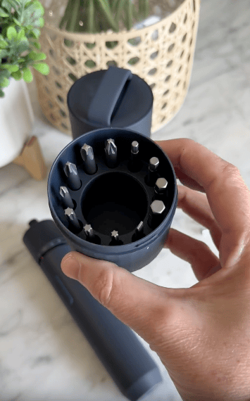 A hand holds a dark gray container with various interchangeable screwdriver bits, with the main screwdriver and another container on a marble surface in the background.