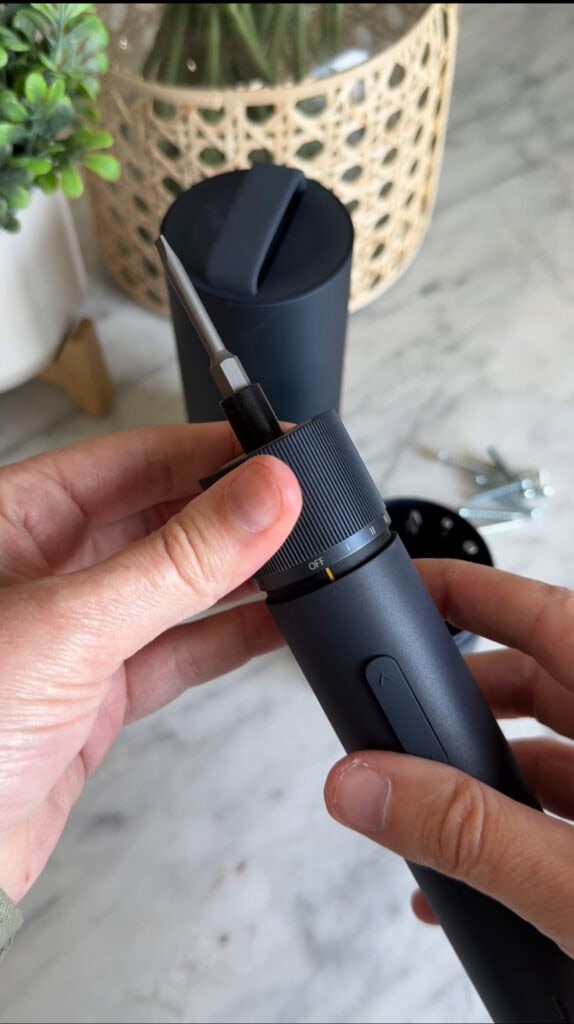 A person holds a black electric screwdriver, adjusting its settings. Screws and a cup are visible on a marble countertop in the background.