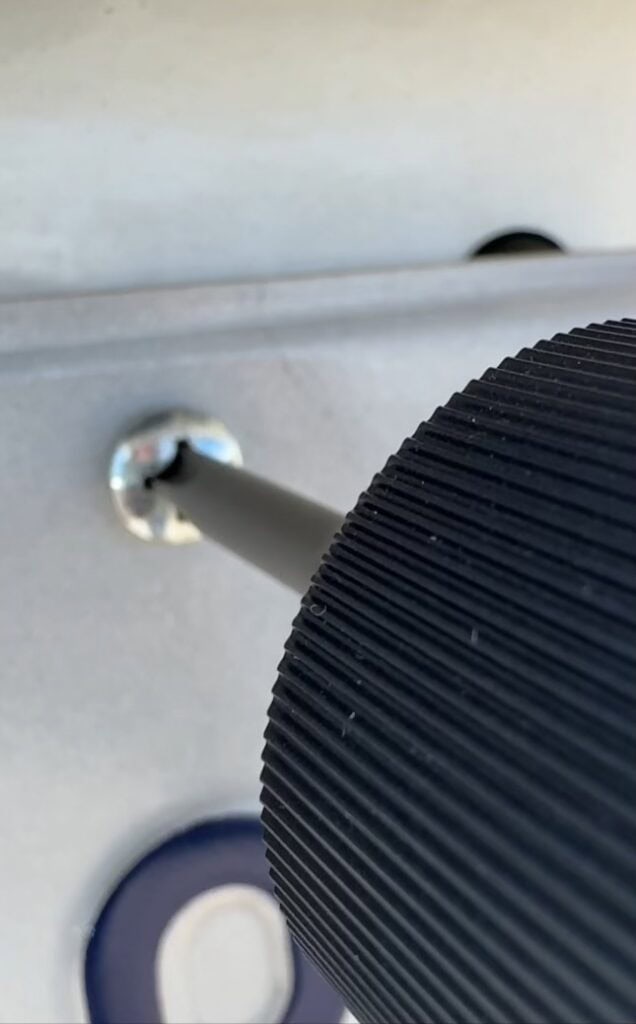 A close-up of a black-handled screwdriver tightening or loosening a screw on a metallic surface, with part of a blue letter visible below.